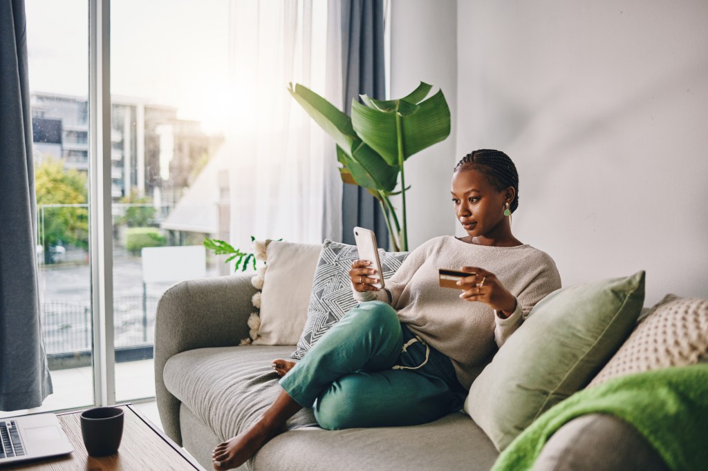 Shot of a young woman using her card and phone to shop online at home